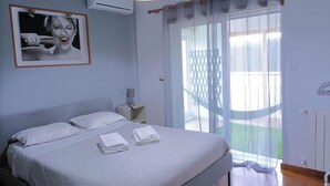 A bedroom with a bed, a nightstand, an air conditioning unit, a window with sheer curtains, and a view of the outdoors. - Welcome to SINTRA HOME, a haven between green hills and the blue Atlantic. (São João das Lampas)