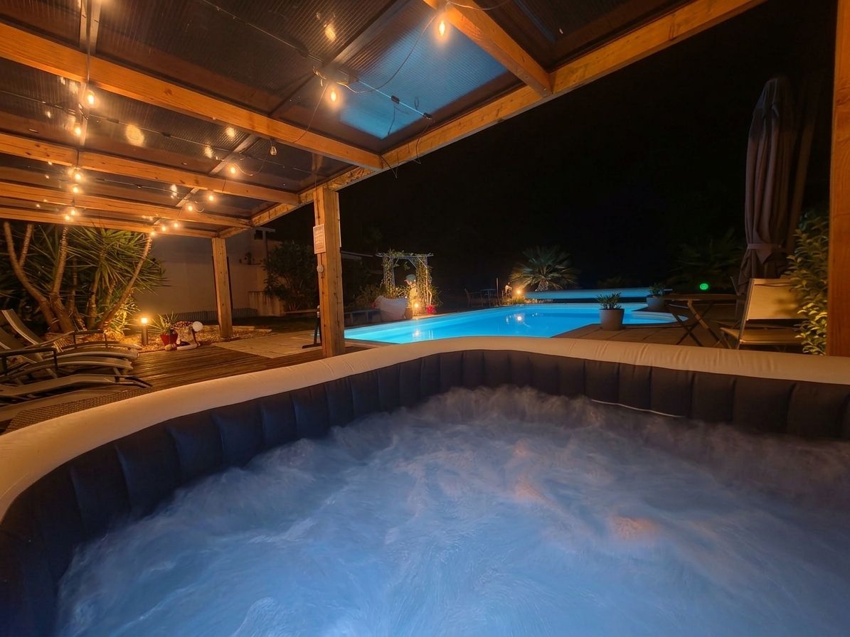 Ett bubbelbad med ett trädäck och ljusslingor, en pool och ett täckt område med växter.