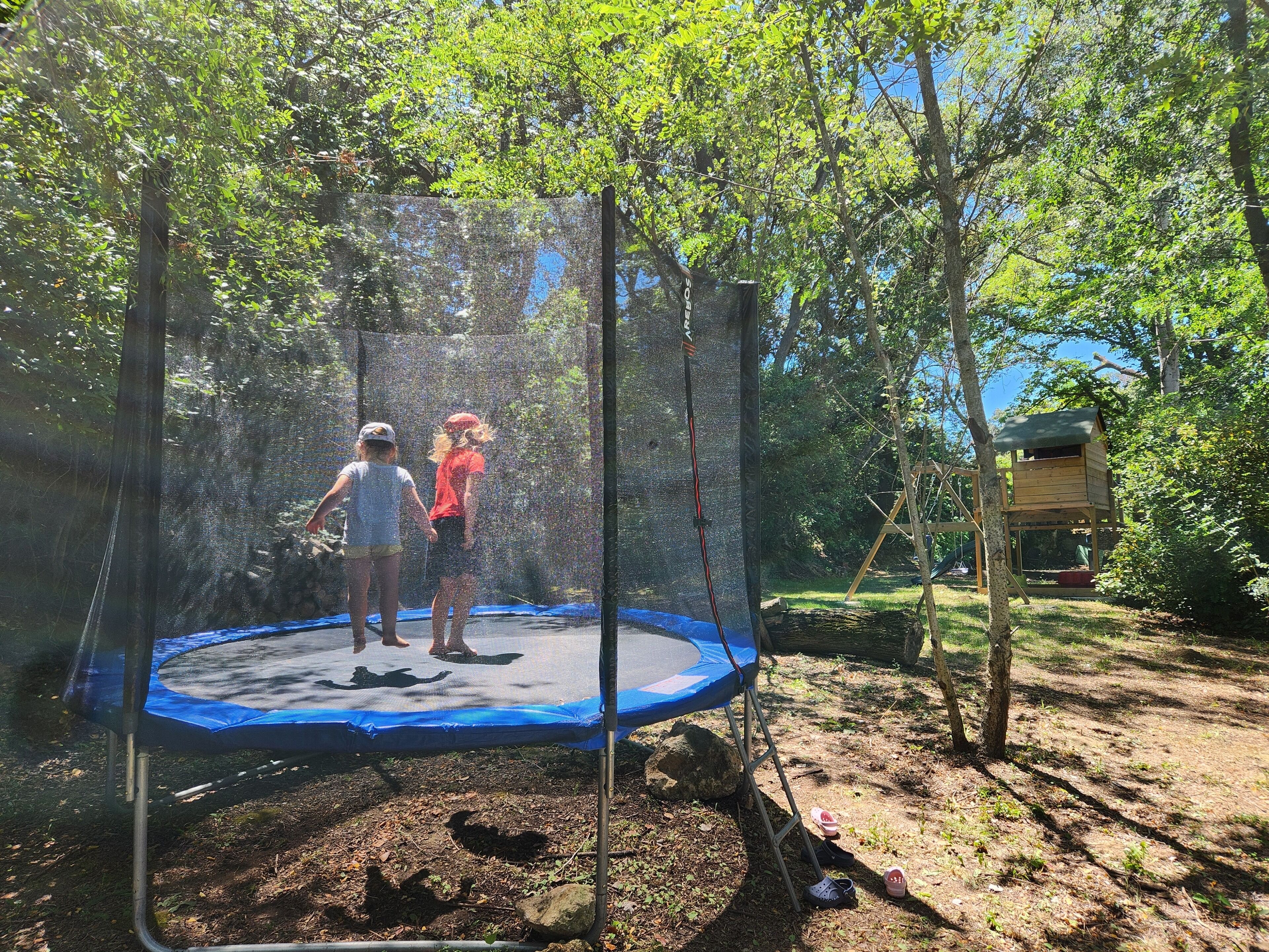Een trampoline in een bebost gebied, met een houten speelhuis op de achtergrond.