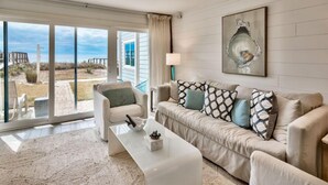 Un salon avec un canapé beige, une table basse blanche et une grande fenêtre donnant sur la plage.