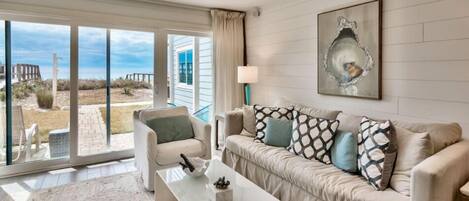A living room with a beige sofa, a white coffee table, and a large window offering a view of the beach.