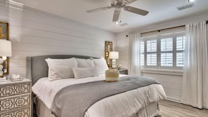 Une chambre à coucher avec un grand lit, un ventilateur de plafond, une table de chevet avec une lampe et une fenêtre avec des rideaux.