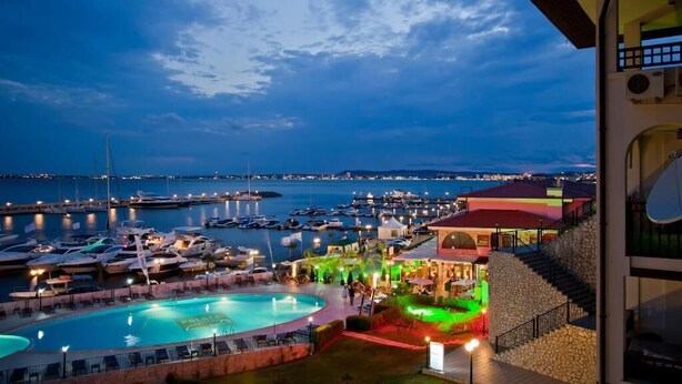 A marina at dusk with boats docked, a swimming pool, and a building with a red roof. - Porto Paradiso Apartments (Sveti Vlas)