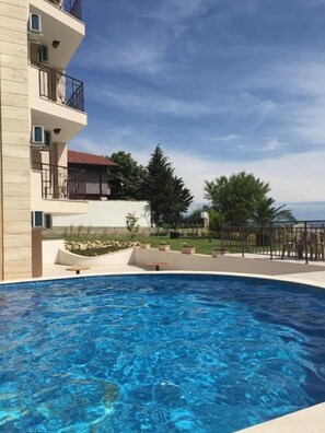 A swimming pool with a clear blue water, surrounded by a building with balconies and a view of trees and a clear sky. - Porto Paradiso Apartments (Sveti Vlas)