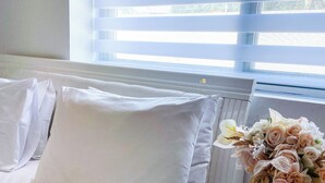 A hotel room with a bed, white bedding, a vase with flowers, and a window with blinds.