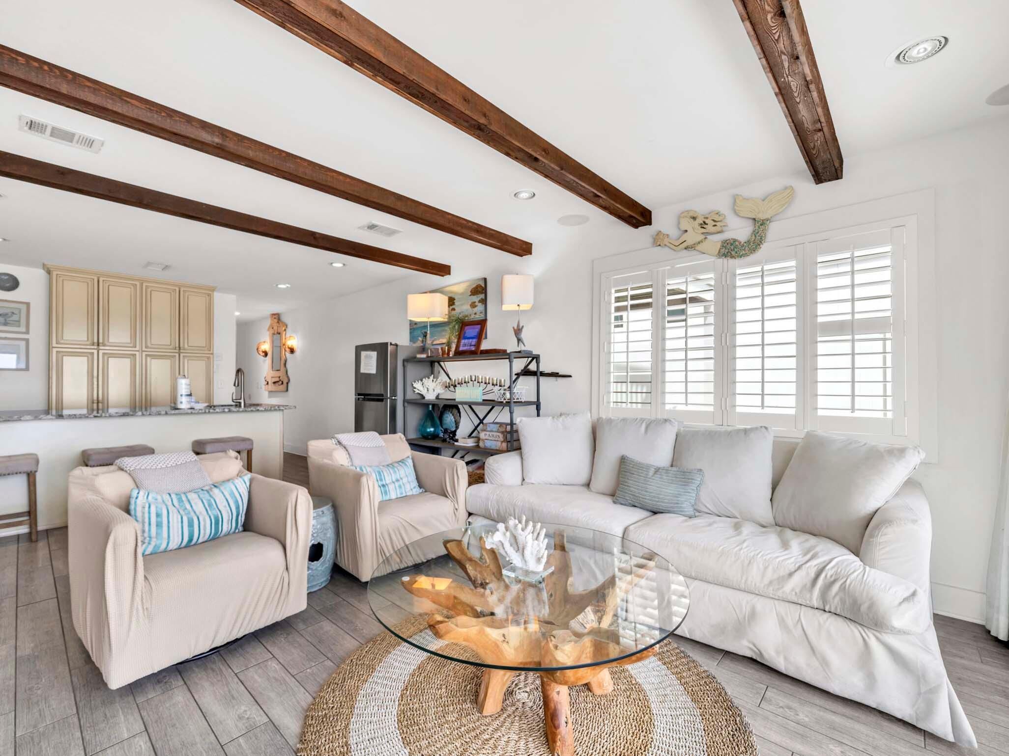 A spacious living room with a large beige sofa, two armchairs, a glass coffee table, and a wooden ceiling beam.