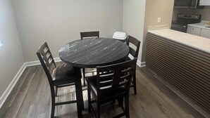 Un coin repas avec une table ronde et six chaises, une cuisine dotée de meubles blancs et d’appareils électroménagers, et un plancher en bois.
