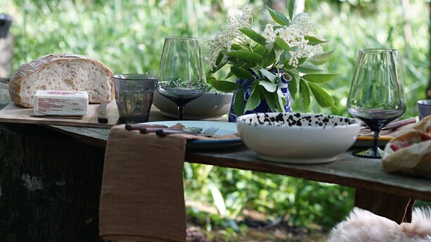 Ein rustikales Essensambiente im Freien mit einem Holztisch, Brot, Weingläsern und einer Vase mit Blumen.