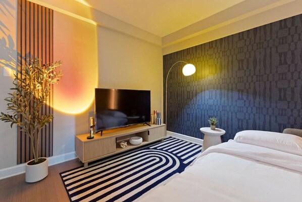 Une chambre d’hôtel moderne dotée d’un téléviseur à écran plat, d’une grande plante en pot, d’un tapis à rayures et d’un mur décoré d’un motif imitant la pierre.