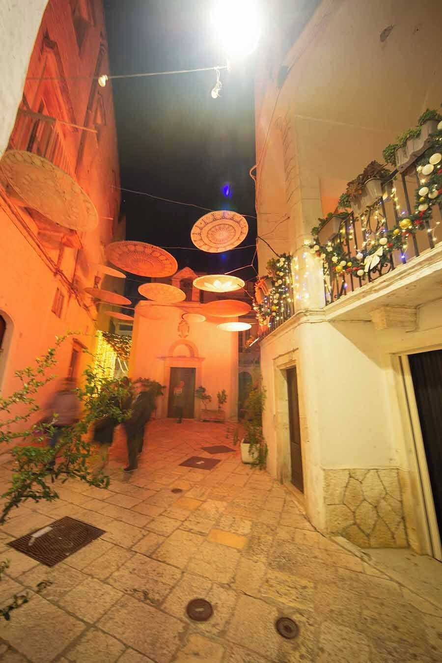 A narrow alleyway with a tiled floor, overhead lights, and a balcony with plants.