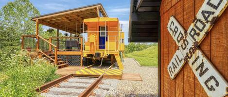A yellow railroad crossing signal structure with a wooden platform and stairs, surrounded by greenery and gravel.