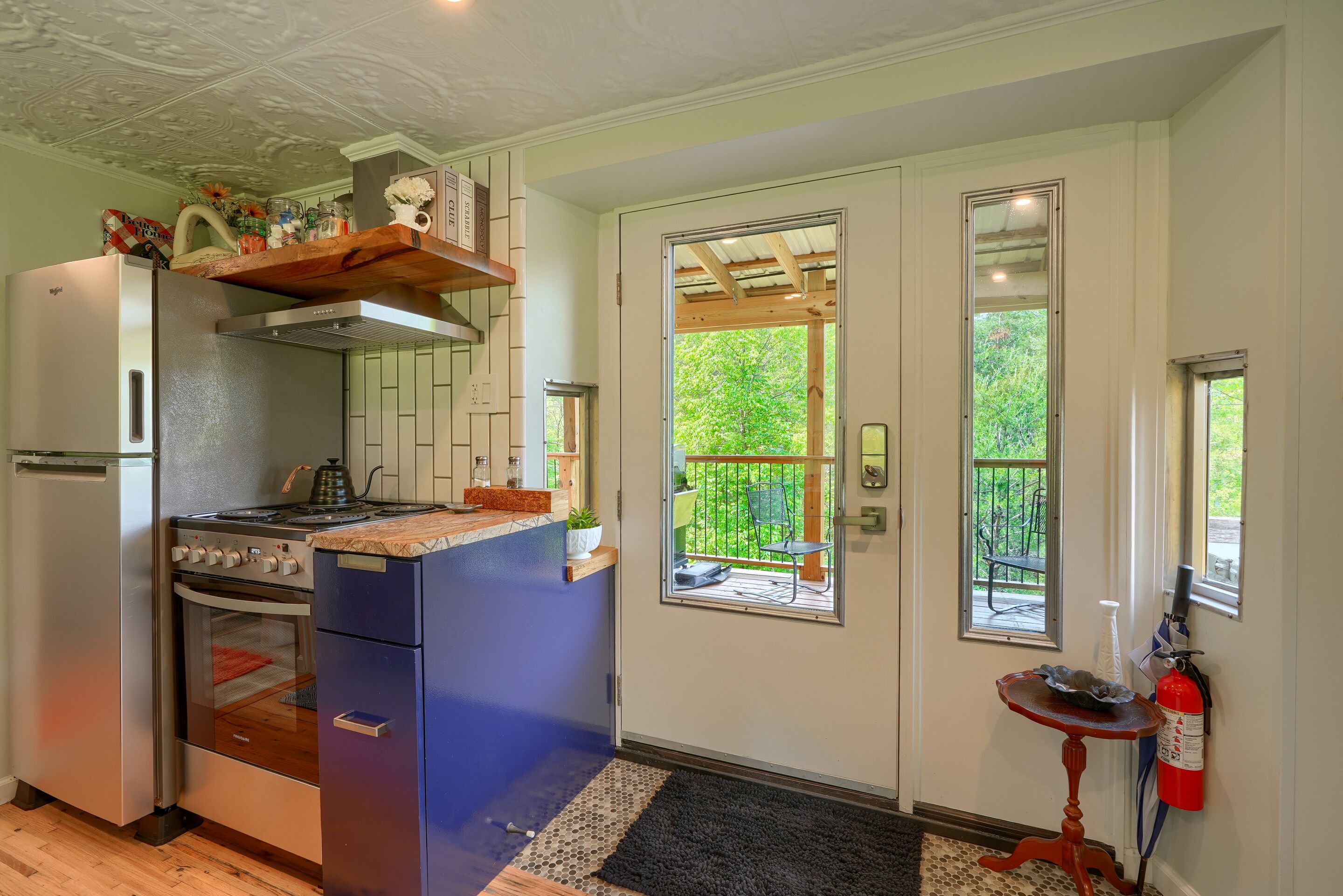 A compact kitchen with a refrigerator, stove, oven, and wooden countertop. There are two windows and a door leading to an outdoor area.
