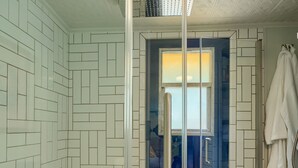 Une salle de bain avec des murs recouverts de carreaux blancs disposés en motif chevron, un tabouret en bois avec des serviettes, et un espace douche doté d’une porte en verre.