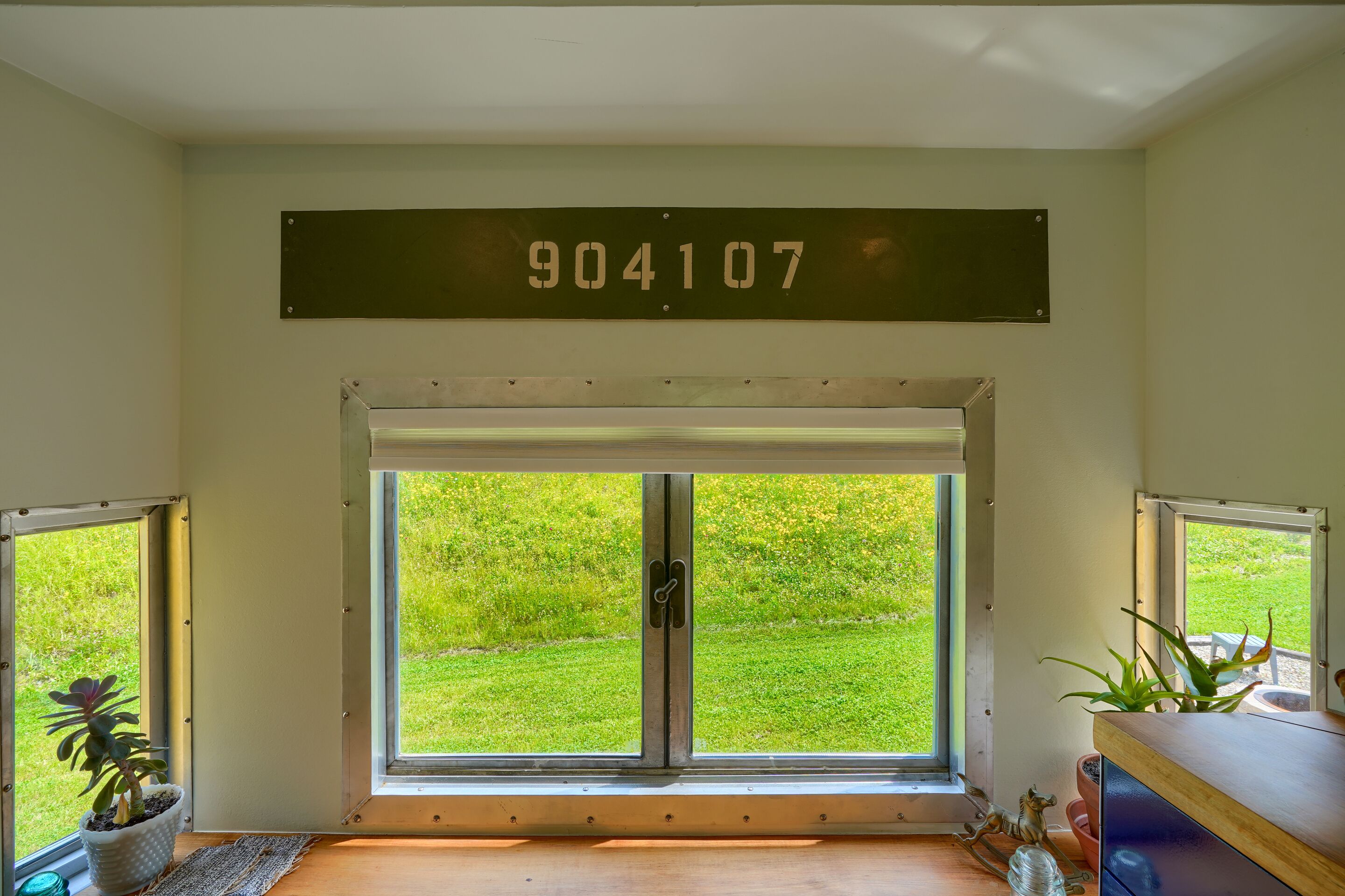 Ett rum med en stor skjutdörr i glas som leder ut till en gräsyta, en metallskylt med numret 904107 och en liten krukväxt på fönsterbrädan.