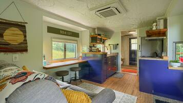 A compact living space with a kitchenette, a sofa with patterned cushions, and a view of greenery outside.