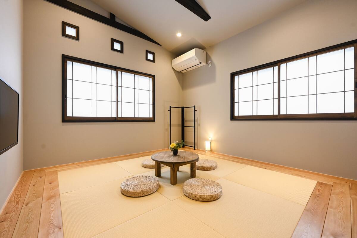 Ein Zimmer mit Tatami-Böden, einem runden Tisch mit Blumen in einer Vase und großen Fenstern mit schwarzen Rahmen.