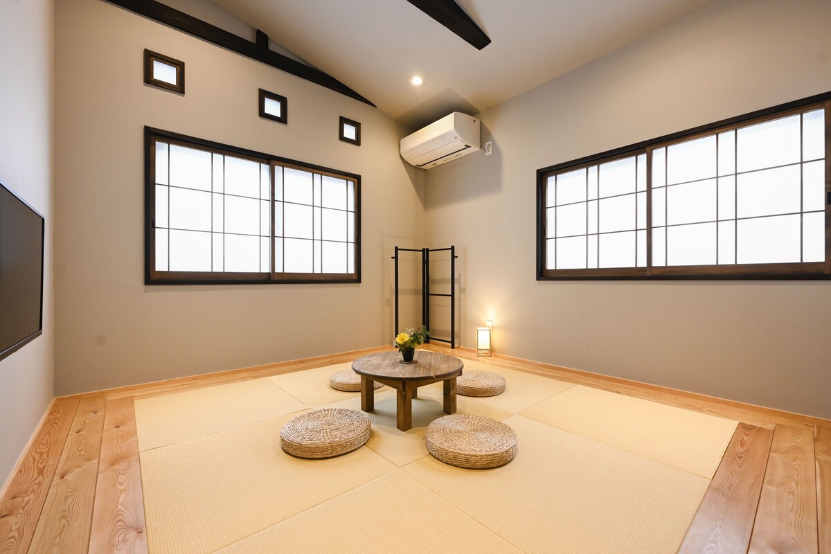 A room with tatami flooring, a round table with a vase of flowers, and large windows with black frames.