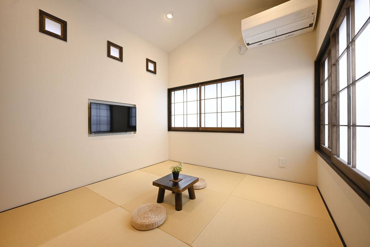 Ein Zimmer mit Tatami-Böden, einem kleinen Tisch mit einer Pflanze, einem Fernseher und einem großen Fenster.