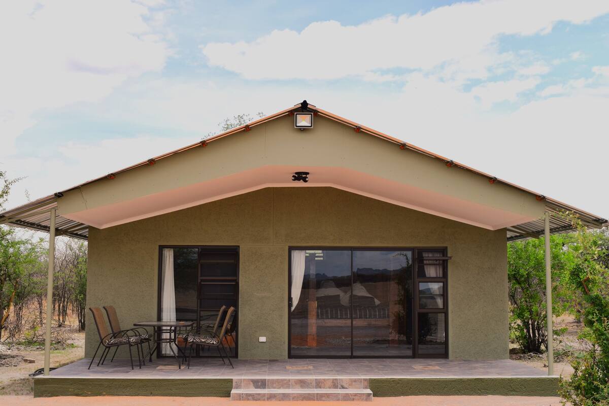 A modern building with a flat roof, large sliding glass doors, and a covered patio area with chairs and a table.