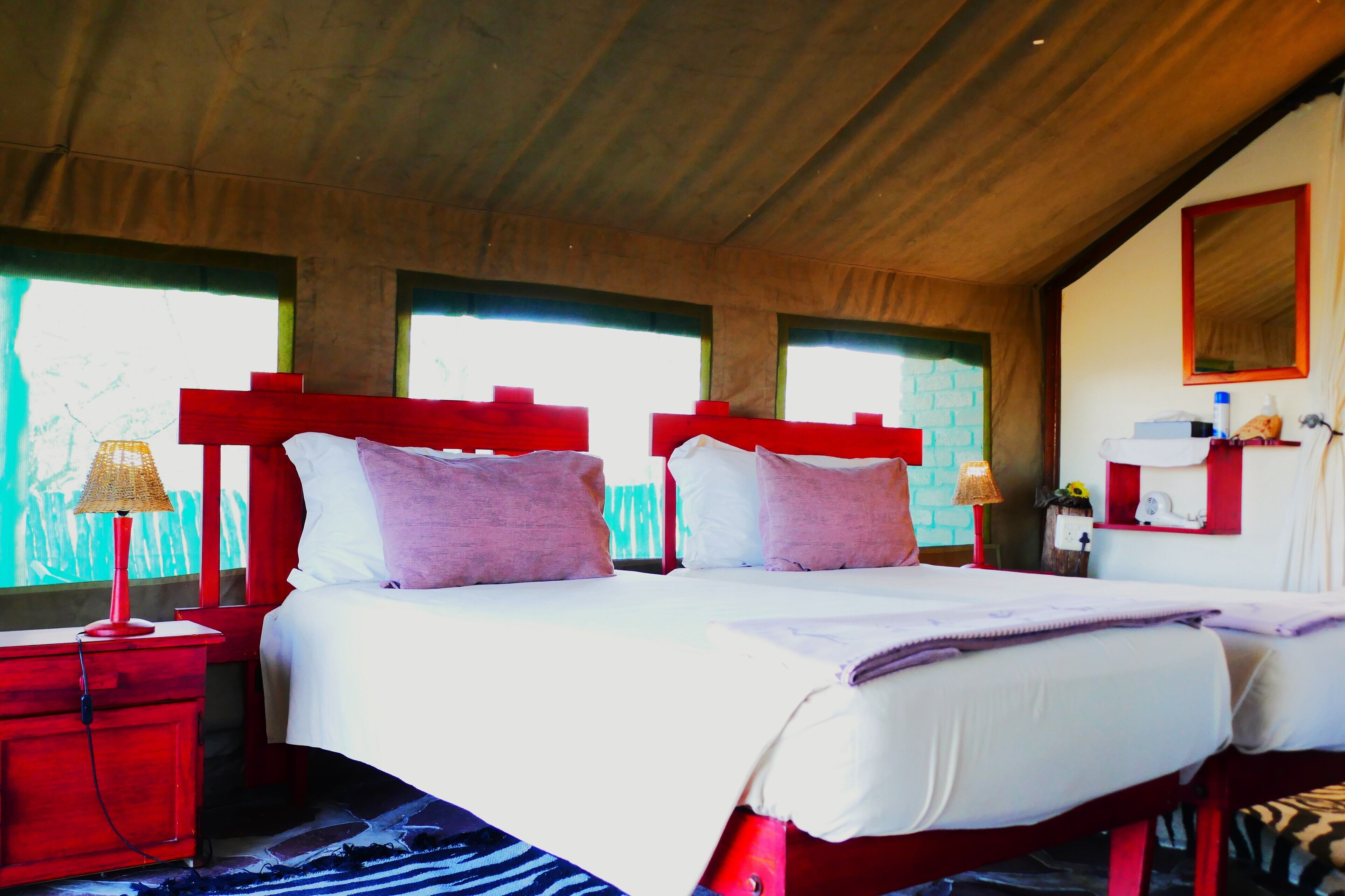A room with two beds, each with red headboards, white bedding, and purple pillows. There are two bedside tables with lamps, a mirror, and a small shelf with items.