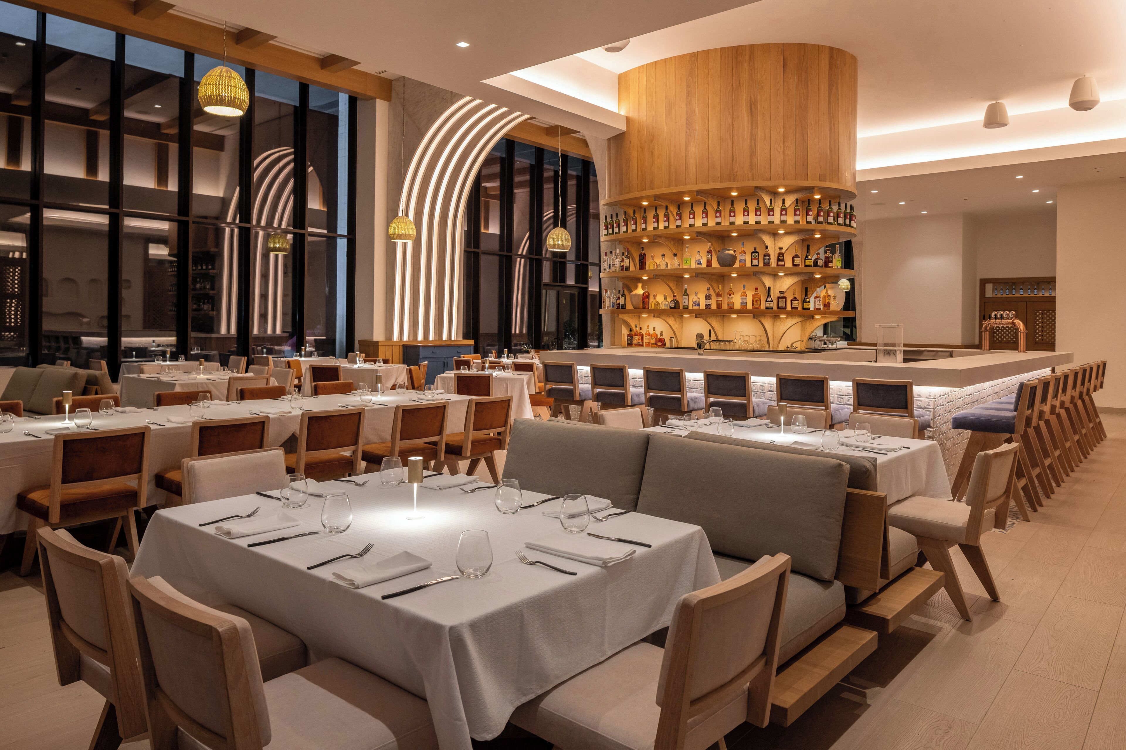 Un restaurant doté de nombreuses tables, d’un bar avec des étagères remplies de bouteilles et d’une grande fenêtre en arc de cercle.