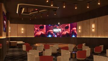 Un espace lounge moderne avec des fauteuils à motifs, un grand écran diffusant des images du « KASSETTE Karaoke Bar » et un éclairage d’ambiance.