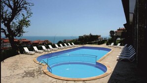 A swimming pool with lounge chairs, a clear view of the sea, and buildings in the background.