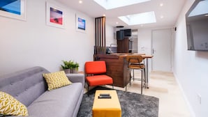 A modern living room with a grey sofa, a red chair, and a small yellow ottoman. There is a skylight above the sofa, a kitchen area in the background, and a grey rug on the floor. - Studio37 - A cosy, stylish central hideaway | Pass the Keys (Cirencester)