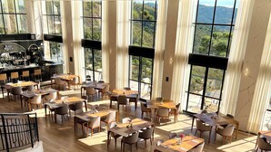 Un'ampia sala da pranzo con numerosi tavoli e sedie, grandi finestre con vista sull'esterno e un'area bar sullo sfondo.