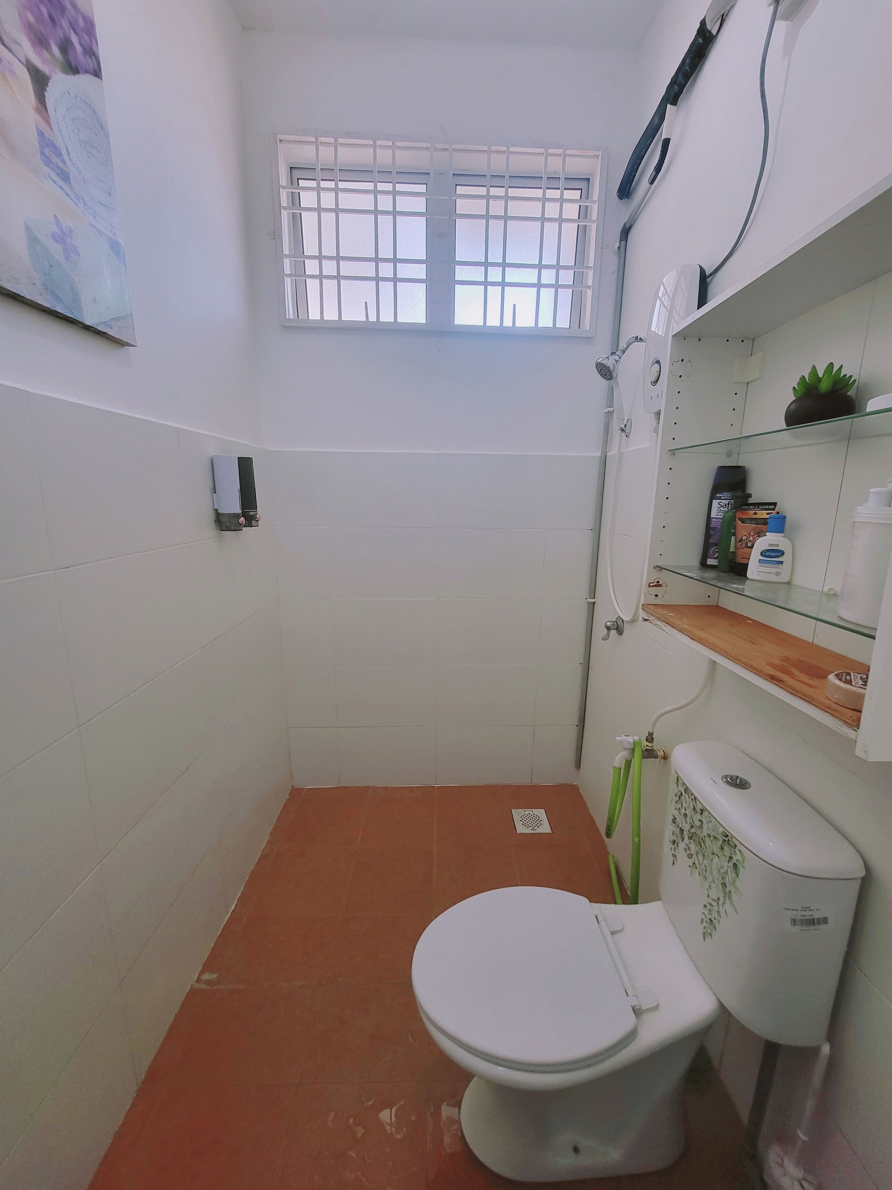 A small bathroom with a toilet, a window, a shower, and shelves with toiletries.