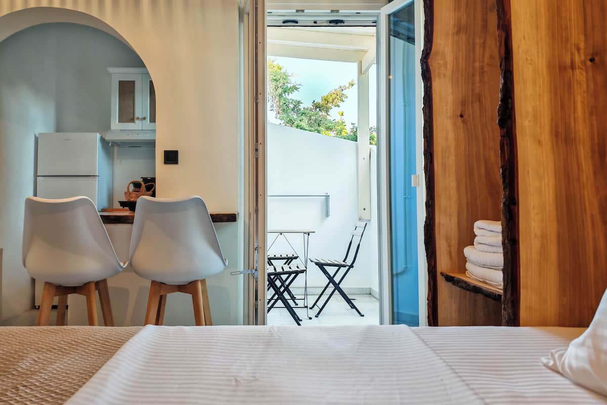 A bedroom with a bed, a desk with two chairs, a kitchen area with a fridge, and an open door leading to an outdoor area with a table and chairs.