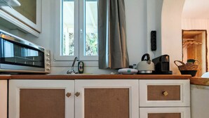 Une cuisine équipée d’un four à micro-ondes, d’une bouilloire et d’une autre cafetière sur le plan de travail. Une fenêtre avec des rideaux offre une vue sur la verdure environnante.