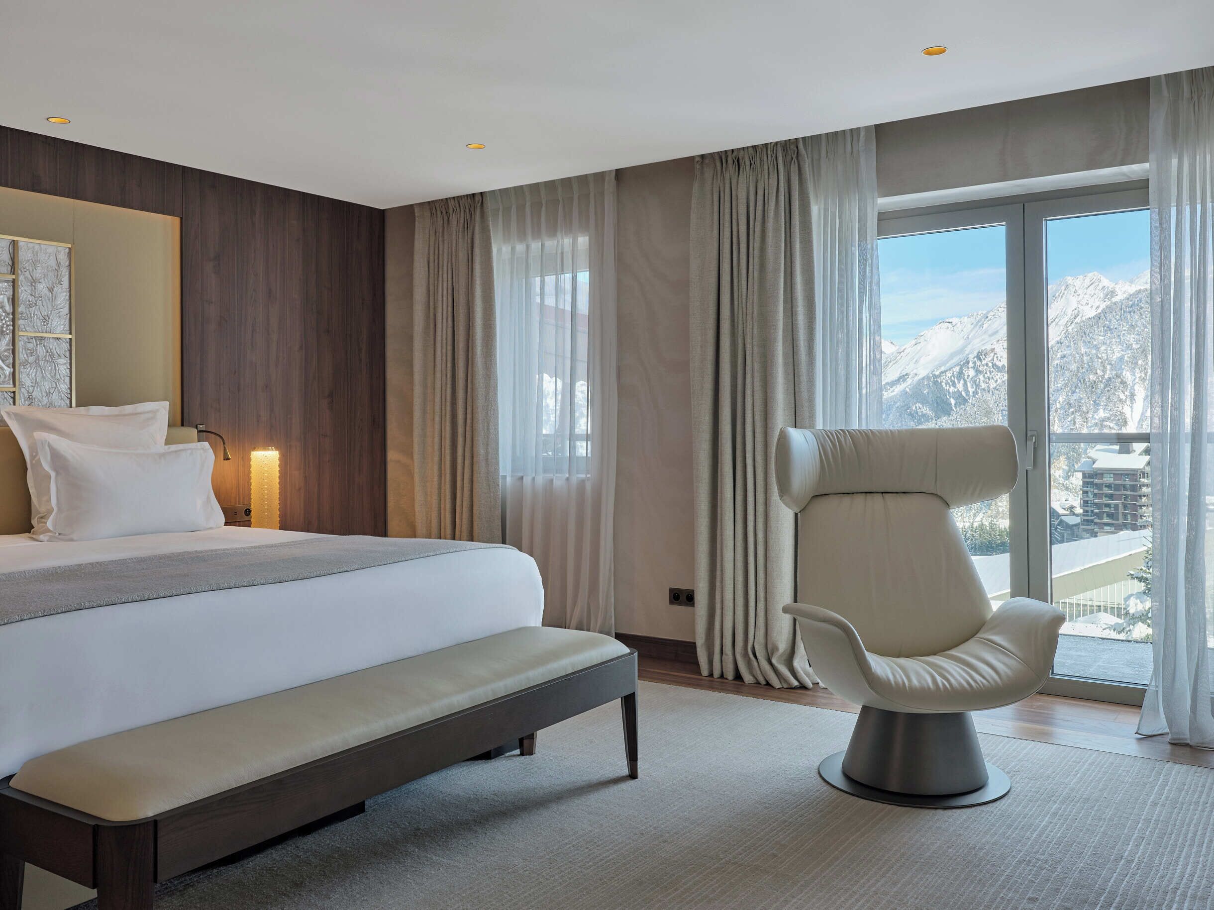 Een hotelkamer met een bed, een bankje, een stoel en een groot raam met uitzicht op de bergen.