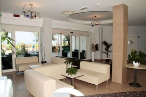 A modern lobby with a beige sofa, a small table with plants, and large windows offering a view of the outdoors. - Bellevue (Cattolica)