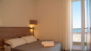 A hotel room with a bed, a nightstand, a wall-mounted lamp, and a view of the beach through a sliding glass door.