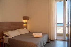 A hotel room with a bed, a nightstand, a wall-mounted lamp, and a view of the beach through a sliding glass door. - Bellevue (Cattolica)