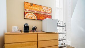 Een houten dressoir met een koffiezetapparaat, waterkoker en een witte koelkast. Daarboven hangt een kleurrijk abstract schilderij aan de muur.