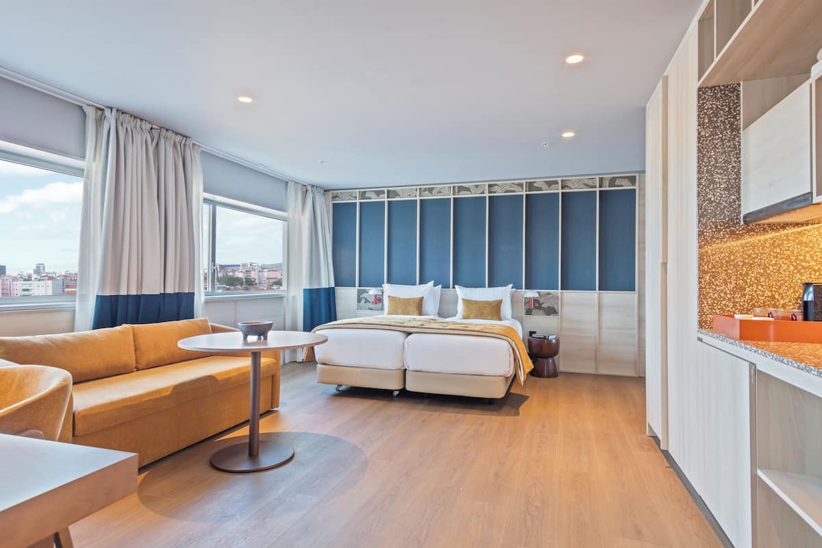 A modern hotel room with a large bed, a sofa, a small table, and a kitchenette.