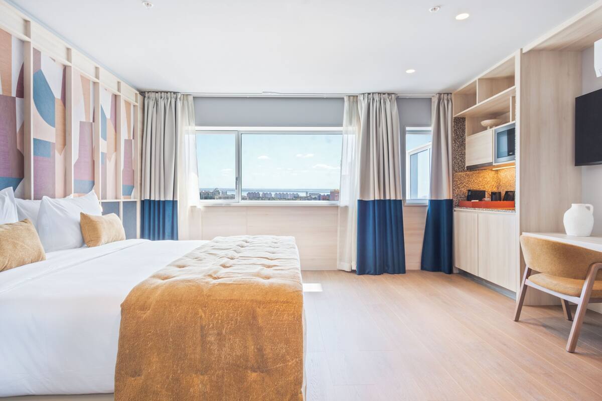 A modern hotel room with a large bed, a desk, and a view of the city.