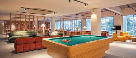 A modern indoor space with a pool table, sofas, and a large window providing natural light.