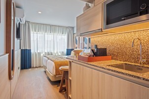 A compact hotel room with a kitchenette, a bed with a headboard, a small stool, and a window with curtains.