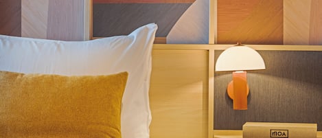A neatly made bed with white and mustard pillows, a wooden headboard, and a wall-mounted lamp with a label.