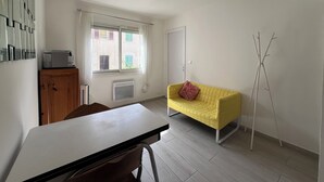 Una stanza con un tavolo bianco, un divano giallo, un armadio in legno, un termosifone, una finestra con tende e un appendiabiti.