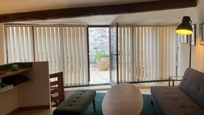 A cozy living room with a sofa, a bench, and a table. There is a large window with blinds, a lamp, and a view of the outside.