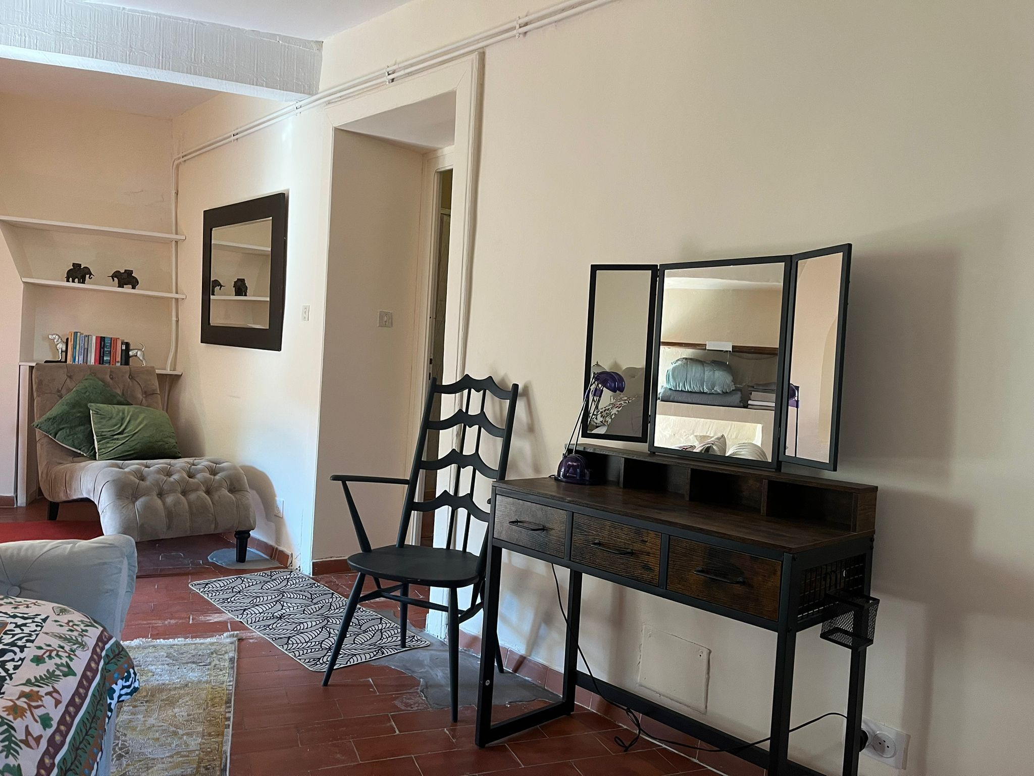Een kamer met een stoel, een klein tafeltje met lades, een spiegel en een plank met boeken en decoratieve objecten.