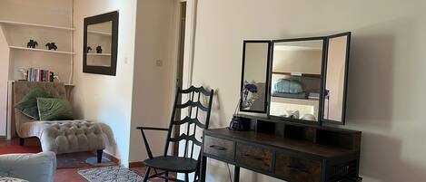 Een kamer met een stoel, een klein tafeltje met lades, een spiegel en een plank met boeken en decoratieve objecten.