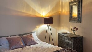 A bedroom with a wooden ceiling, a bed with a dark cover, a bedside table with a vase, and a floor lamp.