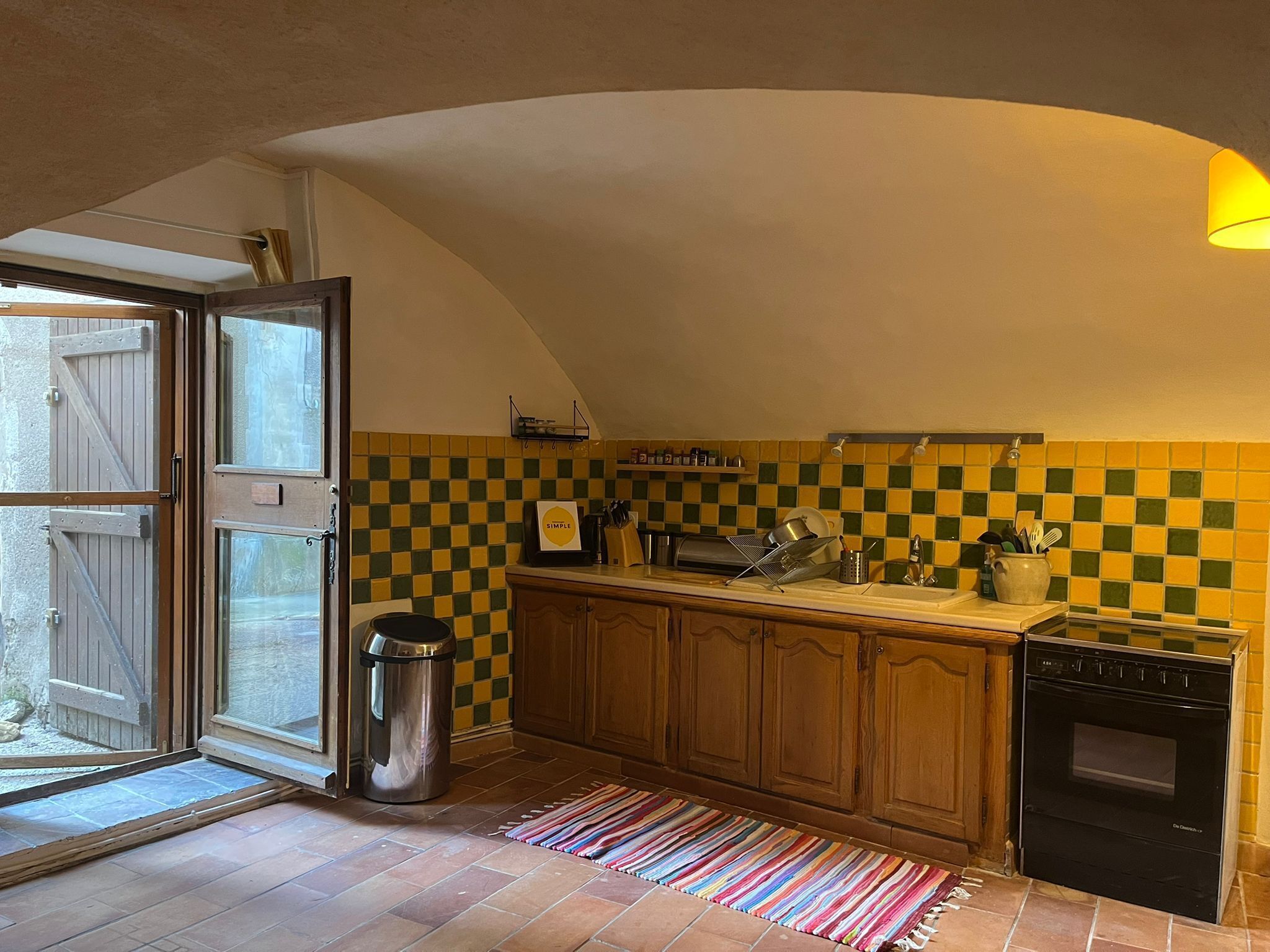 Une cuisine avec un crédence à carreaux, des armoires en bois, une poubelle en acier inoxydable, un tapis coloré et un four noir.