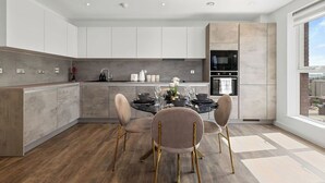 A modern kitchen with a dining area, featuring a central island, white cabinetry, and a view through a large window. - The Lasgidi - One Bed (Barking)