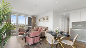 A modern living room with a glass dining table, a sofa, and a view of the city. - The Lasgidi - One Bed (Barking)